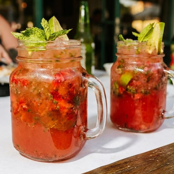 Two strawberry mojitos in mason jars with mint and lime, on a white table.