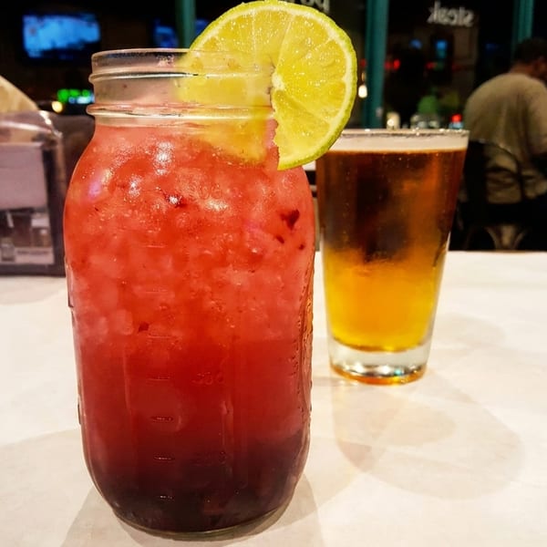 A red drink in a mason jar with a lime wedge and a glass of beer on a table.