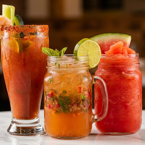 Three vibrant drinks: a Bloody Mary with shrimp, a mojito, and a watermelon slushie on a white table.