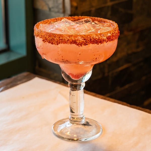 Pink margarita with a chili-salt rim and ice, on a table next to a window.
